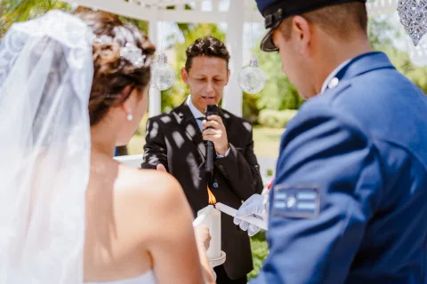 Hombre con traje negro habla por un micrófono frente a una mujer con vestido blanco y un hombre con uniforme azul, junto a dos velas, una con llama visible.