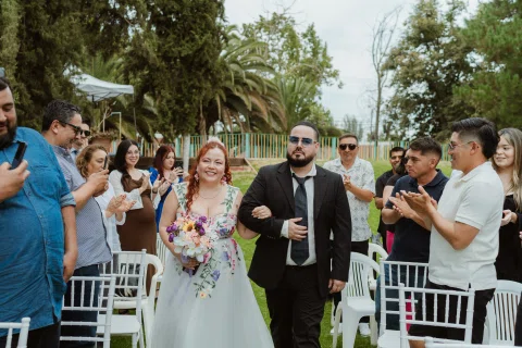 Hombre con traje negro y mujer con ramo caminan entre filas de sillas blancas mientras varias personas aplauden.