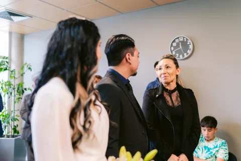 Hombre de traje negro y mujer con vestido blanco permanecen frente a una mujer vestida de negro.