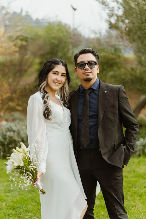 Hombre de traje negro con lentes y mujer con vestido blanco y ramo posan de pie en un parque.