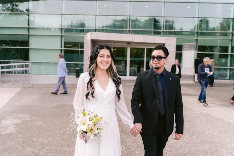 Hombre de traje negro con lentes y mujer con vestido blanco permanecen tomados de la mano frente a un edificio.
