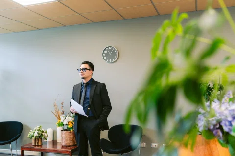 Hombre de traje negro sostiene una hoja junto a una mesa, con un reloj de pared al fondo.