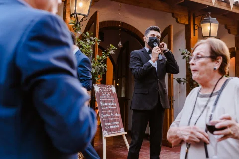 Hombre con traje negro sostiene un teléfono a la altura del rostro en el exterior, mientras una mujer mayor aparece en primer plano.