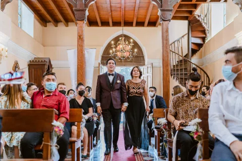 Hombre con traje negro y mujer con vestido negro caminan por el pasillo de la iglesia entre personas sentadas a ambos lados.