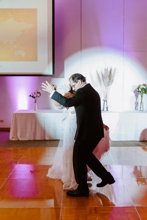 Hombre con traje oscuro baila con una mujer con vestido blanco sobre una pista de madera iluminada con luces de colores.