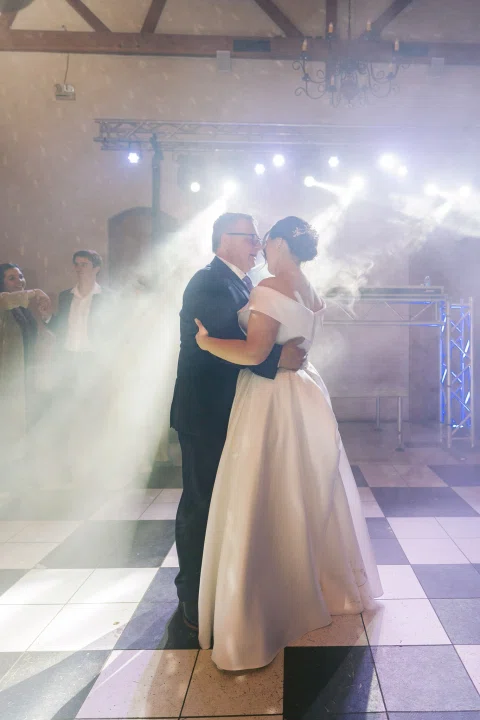 Hombre de traje oscuro baila con una mujer con vestido blanco entre luces y humo.