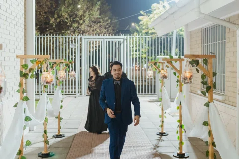 Hombre con traje oscuro camina por un pasillo decorado mientras una mujer vestida de negro aparece detrás.
