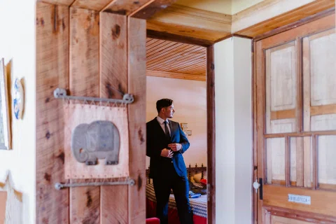Hombre con traje oscuro permanece de pie en una habitación junto a una puerta de madera.