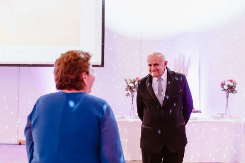 Hombre con traje oscuro permanece frente a una mujer con vestido azul dentro de un salón iluminado.