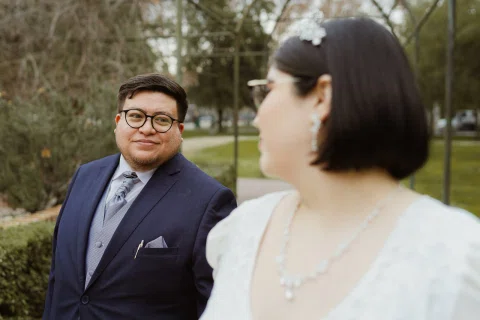 Hombre con traje oscuro y lentes mira hacia una mujer con vestido blanco desenfocada en primer plano.