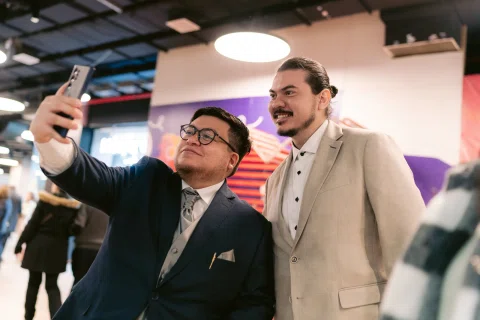 Hombre con traje oscuro y lentes sostiene un celular para una selfie junto a un hombre con traje claro.