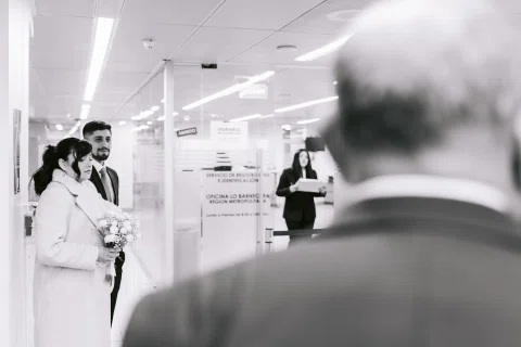 Hombre con traje oscuro y mujer con traje claro permanecen entre varias personas, en una fotografía en blanco y negro.
