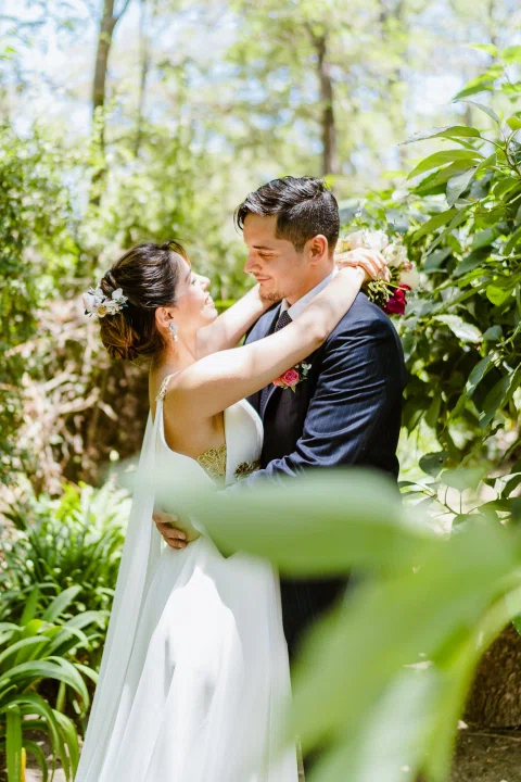 Hombre con traje oscuro y mujer con vestido blanco se abrazan junto a la vegetación.