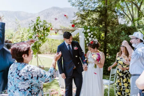 Hombre con traje oscuro y mujer con vestido blanco caminan juntos entre varias personas en el jardín.