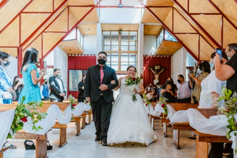 Hombre con traje oscuro y mujer con vestido blanco y ramo caminan por el pasillo de una iglesia entre personas sentadas.