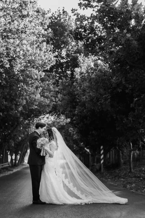 Hombre con traje oscuro y mujer con vestido blanco están de pie juntos en un sendero arbolado, en una fotografía en blanco y negro.