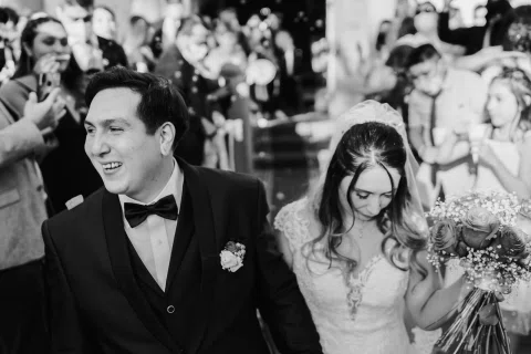 Hombre con traje oscuro sonríe junto a una mujer con vestido blanco y ramo, rodeados por varias personas, en una fotografía en blanco y negro.
