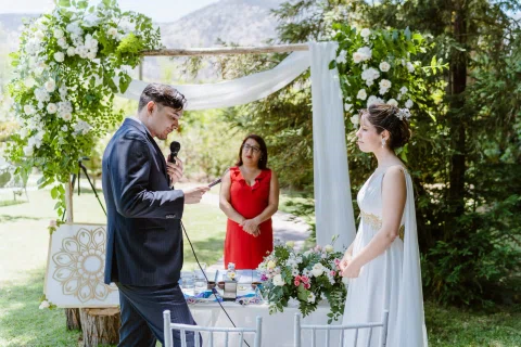 Hombre con traje oscuro y mujer con vestido blanco están de pie frente a una mujer con vestido rojo junto a una mesa con flores.
