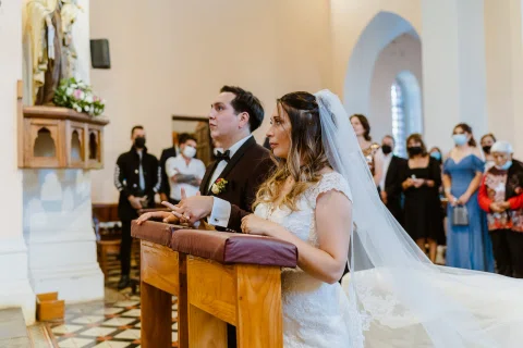 Hombre con traje oscuro y mujer con vestido blanco están de pie junto a un atril dentro de una iglesia.