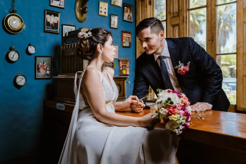 Hombre con traje oscuro y mujer con vestido blanco están juntos junto a una barra de madera con un ramo sobre la superficie.
