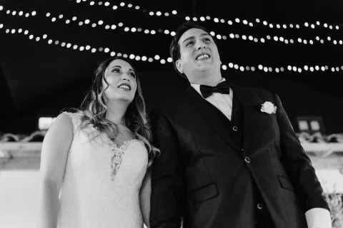 Hombre con traje oscuro y mujer con vestido blanco miran hacia arriba bajo guirnaldas de luces, en una fotografía en blanco y negro.