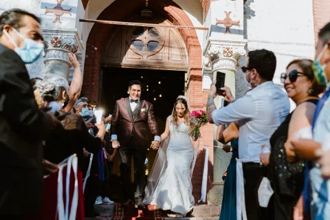 Hombre con traje oscuro y mujer con vestido blanco salen de una iglesia entre varias personas.