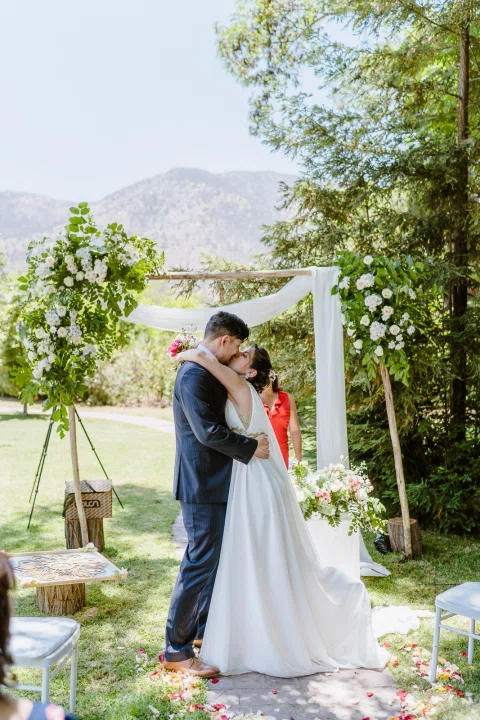 Hombre con traje oscuro y mujer con vestido blanco se besan bajo un arco decorado con flores.