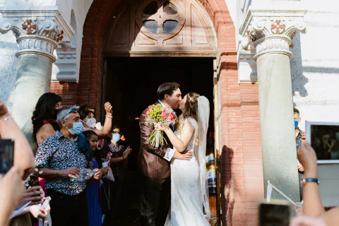 Hombre con traje oscuro y mujer con vestido blanco se besan en la puerta de una iglesia mientras varias personas los rodean.