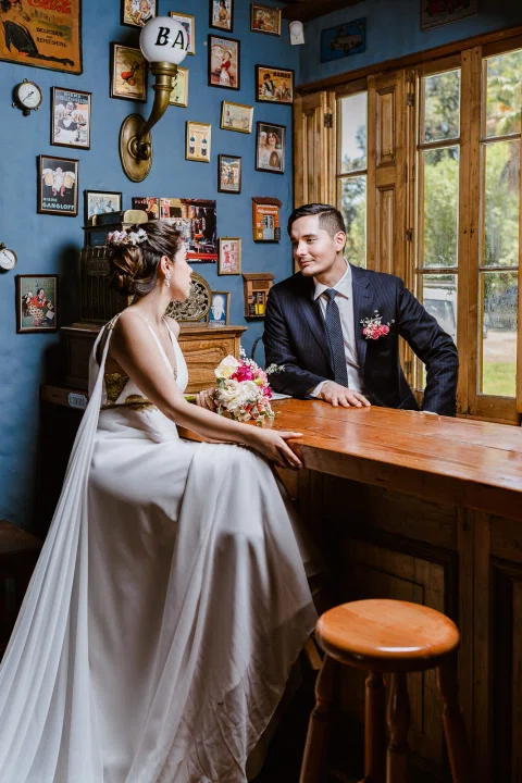 Hombre con traje oscuro y mujer con vestido blanco están sentados junto a una barra de madera en un interior.