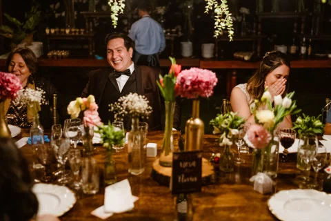 Hombre con traje oscuro y mujer con vestido blanco están sentados en una mesa decorada con flores y velas.