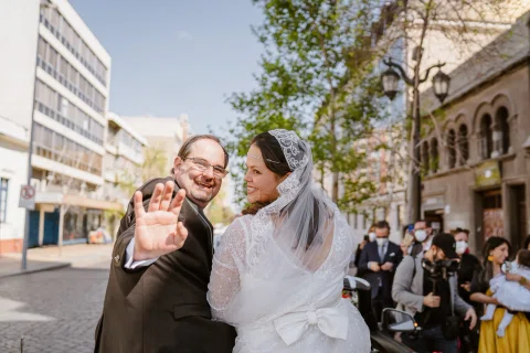Hombre con traje oscuro sonríe y levanta una mano hacia la cámara junto a una mujer con vestido blanco en el exterior de una iglesia.