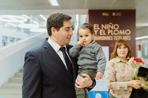 Hombre con traje oscuro sostiene en brazos a un niño pequeño dentro de un espacio interior.