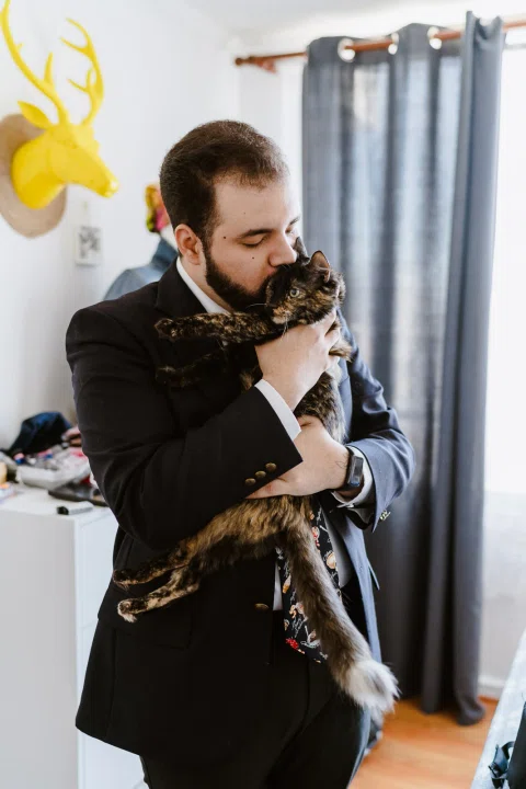 Hombre con traje oscuro sostiene un gato en brazos y lo besa dentro de una habitación.
