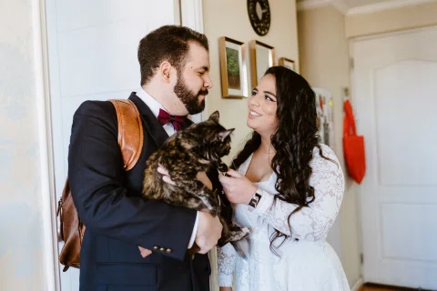 Hombre con traje oscuro y mujer con prenda blanca sostienen juntos a un gato dentro de una habitación.
