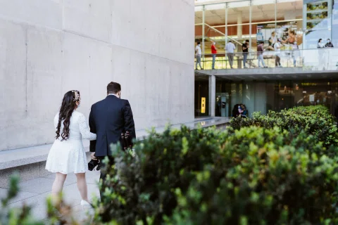 Hombre con traje oscuro y mujer con vestido blanco caminan junto a un edificio de hormigón y vegetación.