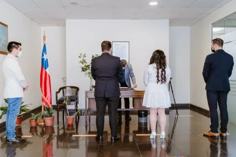 Hombre con traje oscuro y mujer con vestido blanco vistos de espaldas frente a una mesa, con otras personas a los lados.