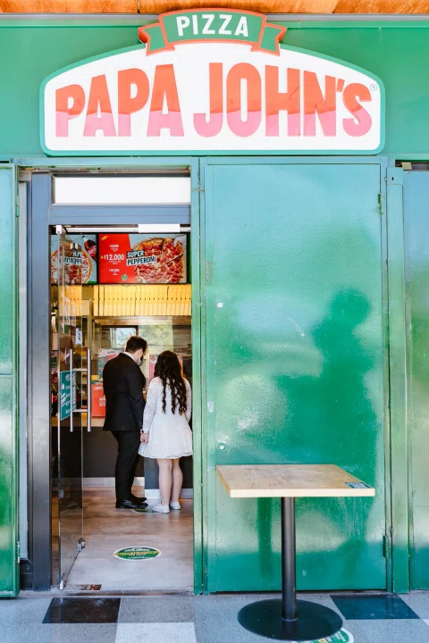 Hombre con traje oscuro y mujer con vestido blanco permanecen en la entrada de una pizzería.