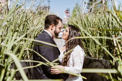 Hombre con traje oscuro y mujer con vestido blanco permanecen frente a frente entre pastos altos.