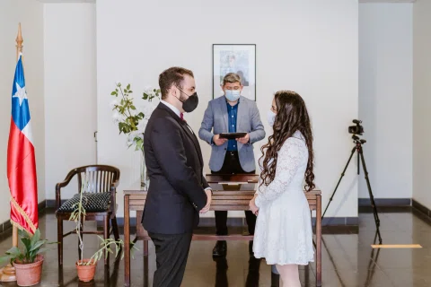 Hombre con traje oscuro y mujer con vestido blanco permanecen frente a una mesa junto a una bandera de Chile.