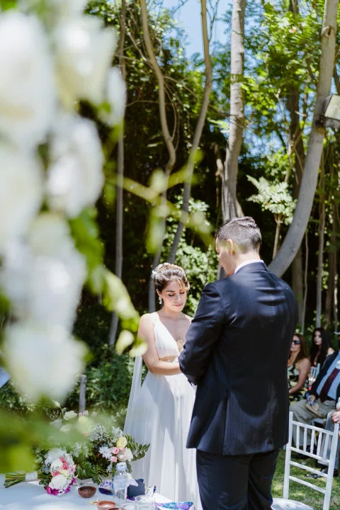 Hombre con traje oscuro y mujer con vestido blanco permanecen de pie frente a la mesa de ceremonia en el jardín.