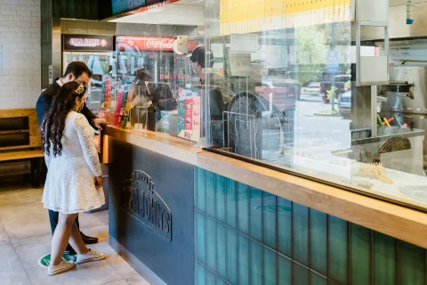 Hombre con traje oscuro y mujer con vestido blanco permanecen frente al mostrador de una pizzería.