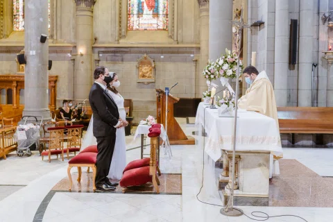 Hombre con traje oscuro y mujer con vestido blanco permanecen frente a una persona con vestimenta religiosa junto al altar de una iglesia.