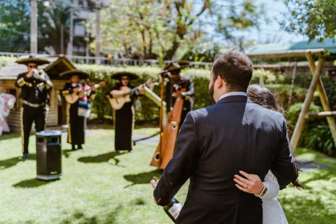 Hombre con traje oscuro y mujer con vestido blanco permanecen abrazados de espaldas frente a varias personas con sombreros e instrumentos en un jardín.