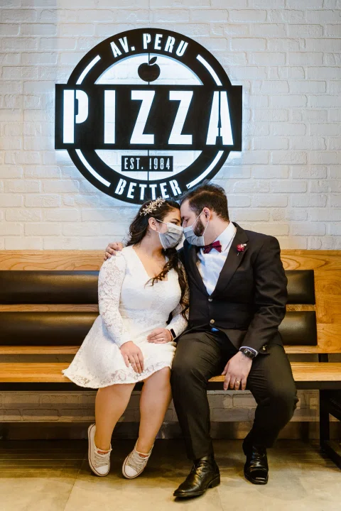 Hombre con traje oscuro y mujer con vestido blanco están sentados juntos bajo un gran letrero circular de pizza.