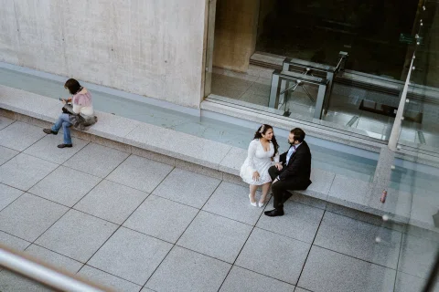 Hombre con traje oscuro y mujer con vestido blanco permanecen sentados en el exterior de un edificio, fotografiados desde un ángulo alto.
