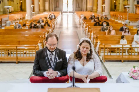Hombre con traje oscuro y mujer con vestido blanco permanecen sentados con las manos juntas dentro de una iglesia.
