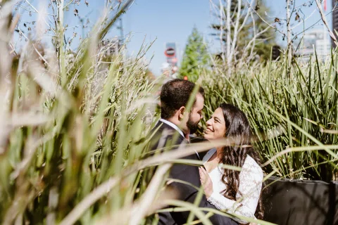 Hombre con traje oscuro y mujer con vestido blanco se miran sonriendo entre pastos altos.