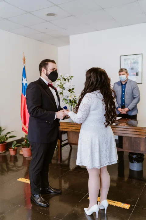 Hombre con traje oscuro y mujer con vestido blanco permanecen tomados de las manos frente a una mesa, con otra persona detrás.