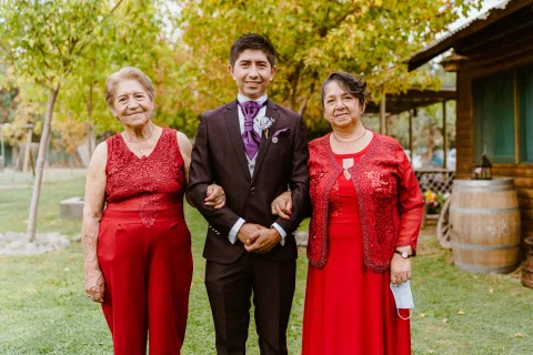 Hombre con traje oscuro posa entre dos mujeres vestidas de rojo en el jardín.