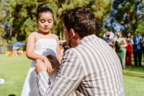 Hombre con traje rayado está arrodillado frente a una niña de vestido blanco en el jardín.